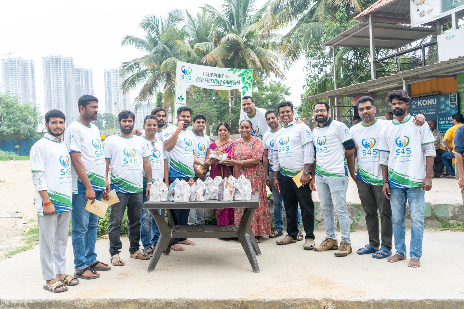 Service4Society volunteers and residents standing around a table of wrapped clay Ganesha idols beneath an I Support Eco Friendly Ganesha banner