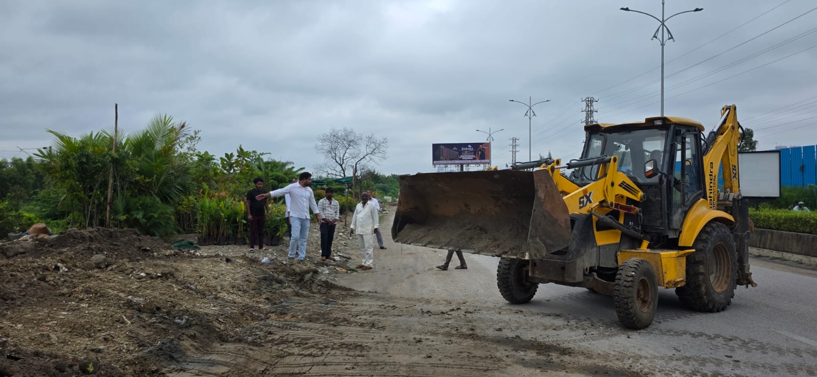 Representatives and residents standing on the RR7 road stretch in Tellapur while an excavator levels the damaged surface